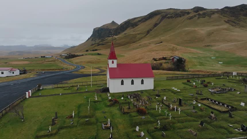 An aerial shot circles a white church with a red roof and a graveyard set against a mountainous backdrop