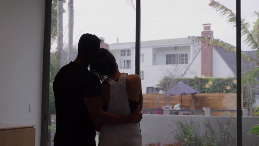A couple is silhouetted as they stand closely together embracing while looking out of large floortoceiling windowsthe view beyond the glass shows a suburban backyard and a white house with a patio area in the distance