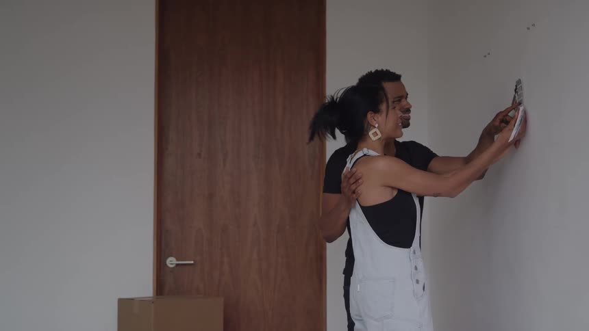 A man stands behind a woman embracing her while she holds a small lightcolored object up to a white wall and they both smile while looking at the placement in a sparsely furnished room with a large wooden door in the background