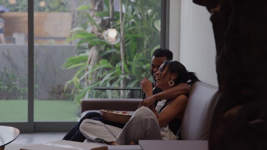 Young man and young woman sitting on a couch hugging and laughing while talking next to a glass window showing a lawn indoors during day time