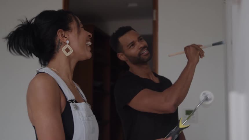 A couple enjoys painting a building wall with white paint at midday joking and laughing together as they work