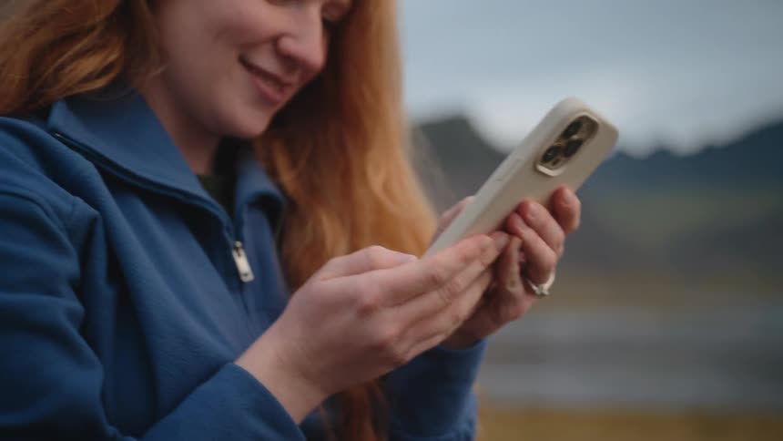 A smiling woman with long red hair and a blue fleece jacket uses a smartphone outdoors in a natural overcast environment