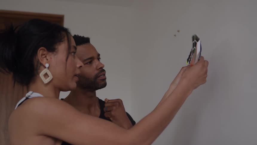 A man and a woman are standing close together looking intently at a wall in their homethe woman is holding up a paint swatch deck comparing various color chips to the current wall color as they discuss home decorating or renovation plans