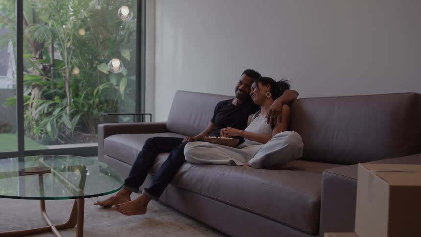 This video features a couple relaxing on a couch in a modern living roomthey are sitting close with the mans arm around the woman watching television and eating popcorn in a room with large windows overlooking a garden