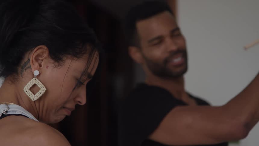 A woman with her hair pulled back and wearing large patterned earrings is seen smiling and interacting with a man standing slightly behind herthey appear to be engaged in an activity together possibly cooking or preparing something as the man holds