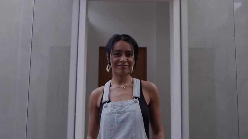 A young woman stands inside a bathroom smiling warmly while wearing a jumpsuit and large square earrings