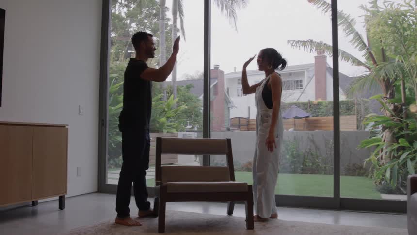A man and a woman are standing on opposite sides of a small wooden chair inside a modern welllit room with large windows overlooking a backyardthey share a highfive over the back of the chair suggesting they have just completed a task or are celebr