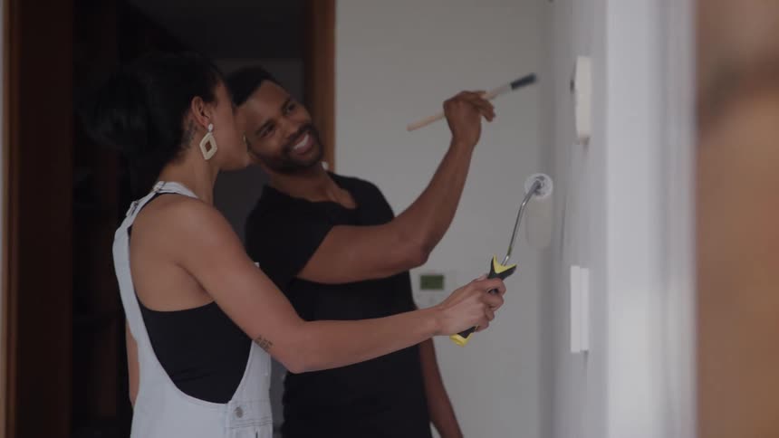 A smiling couple paints an interior wall with a roller and brush in a home renovation