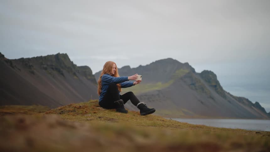 A woman sits on a grassy mound taking a selfie with mountains and a still lake behind her