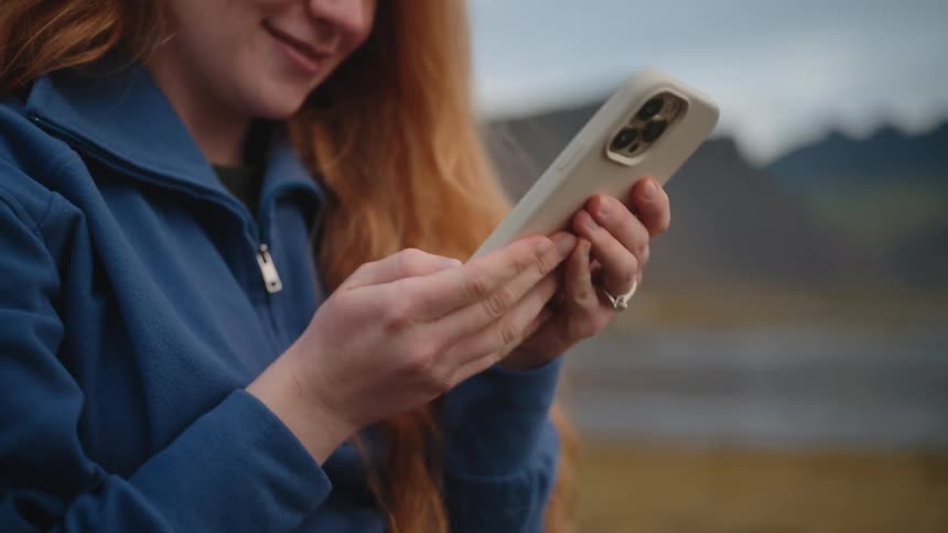A smiling woman with long red hair and a blue fleece jacket uses a smartphone outdoors with a blurred mountain range in the background