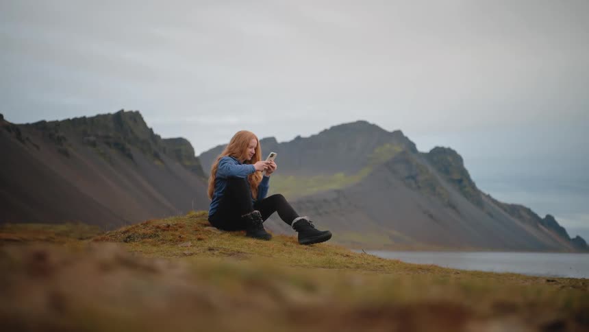 A smiling woman sits on a grassy mound texting on her phone with rocky mountains behind