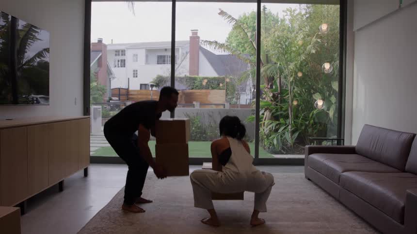 A happy couple moves boxes in a bright living room with large windows and a patio garden