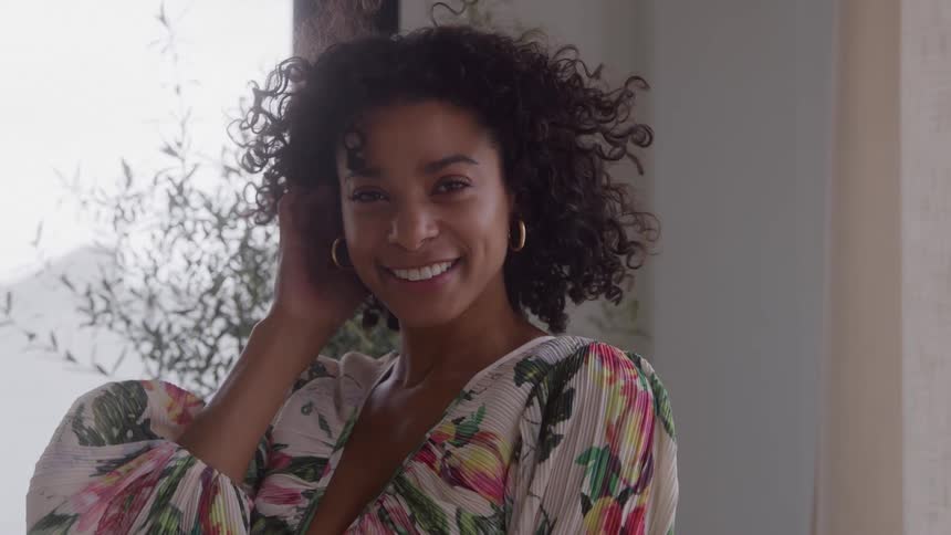 A smiling woman with curly hair sweeps her hair back from her face while looking at the camera