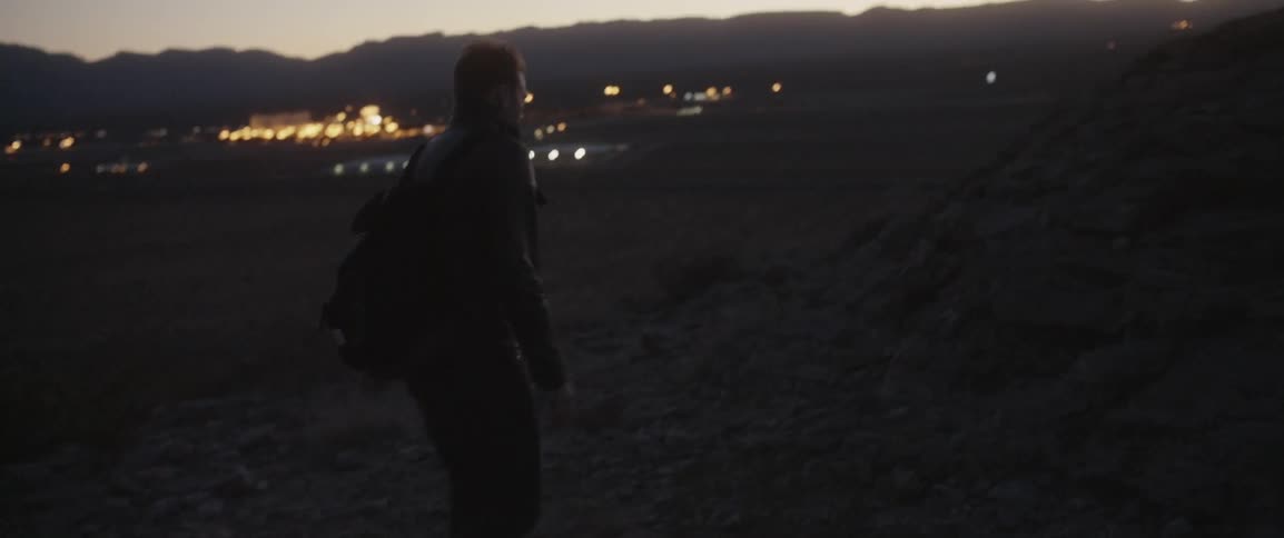 A man walks over a rocky area with building lights in the distance
