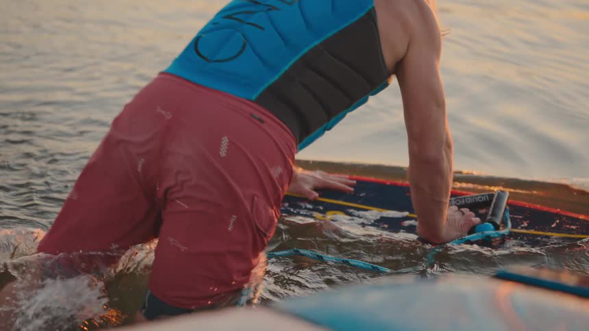 This video is a closeup shot showing a person preparing for a water sport likely wakeboarding or wakesurfingthe person is wearing a blue life vest and red shorts kneeling in the water next to a colorful board gripping the tow handle as water splash