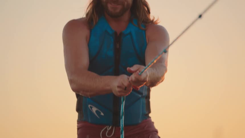 A longhaired man with a beard and a backwards baseball cap smiling as he grips the tow rope while engaging in a water sport in the beach at sunset
