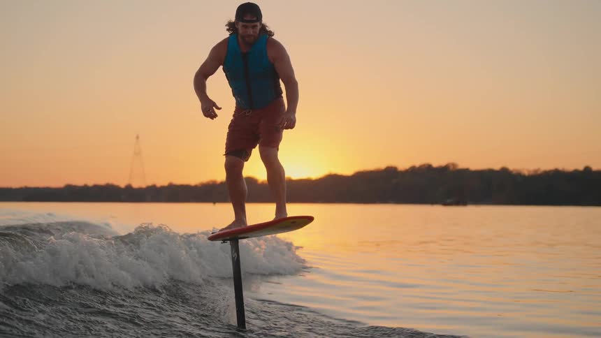 A man wearing a blue life vest and a cap is hydrofoiling on a lake during sunset he is riding on a small wave and the sky behind him is painted in warm orange and yellow hues