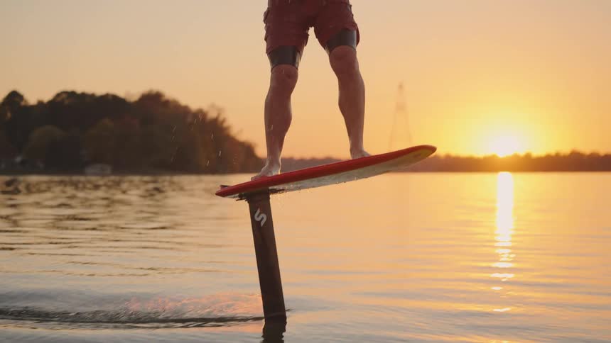 A man rides a hydrofoil board across a lake while being towed during a golden sunset