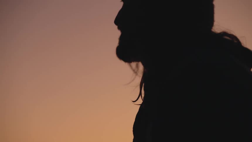 This video begins with a closeup of a metal edge covered in water droplets likely a railing or skateboard then transitions to a silhouette of a persons head and shoulders against a warm orange sunset the person is wearing a beanie and has long hair
