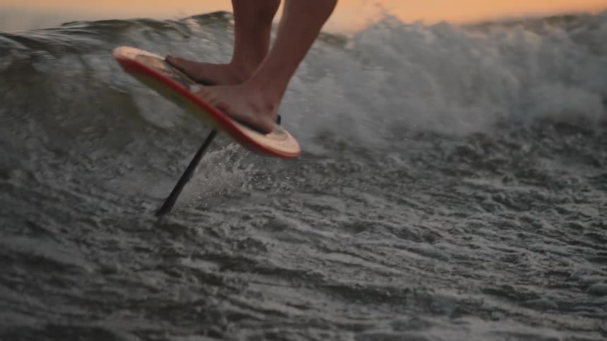 This closeup video shows the bare feet of a person riding an efoil a type of hydrofoil surfboard over choppy water the board which is dark with orange and yellow accents is visibly lifted above the water surface by the hydrofoil mast as it creates a
