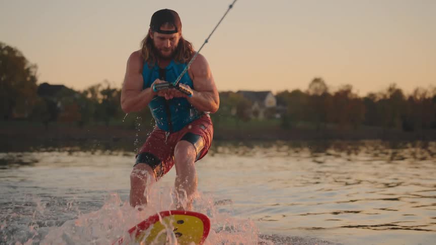 A man with long hair wearing a blue tank top red shorts and a life vest is wakesurfing or wakeboarding on a lakehe is holding onto a tow rope and riding a yellow and red board generating a splash of water with the scene set during golden hour