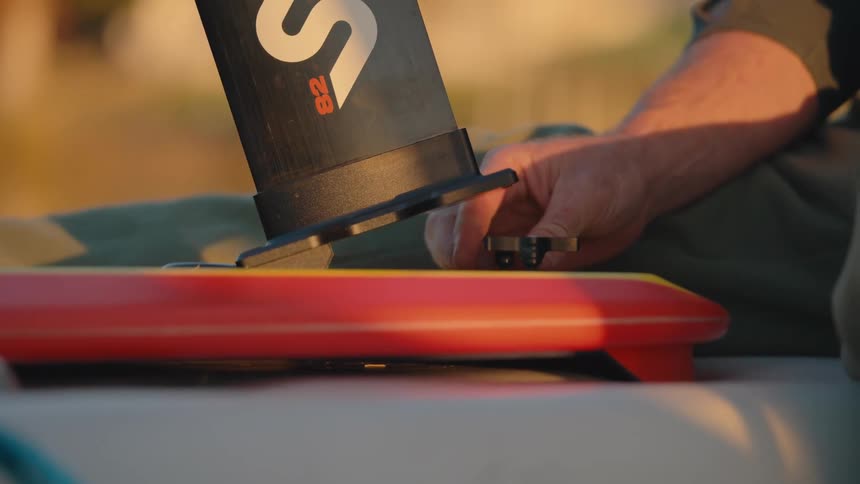 This is a closeup video showing a person assembling a hydrofoil setup attaching the mast to a brightly colored boardthe hands work to tighten the screws connecting the black labeled mast to the red and yellow underside of the board in soft golden l