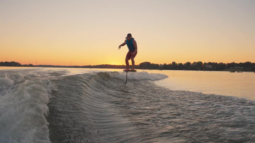 A person foil surfs on the wake behind a boat during a vivid golden sunset