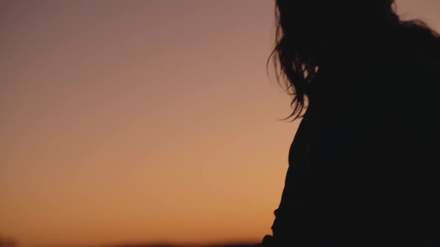 A silhouetted woman stands holding a surfboard with water droplets on it against the warm orange glow of a sunset sky