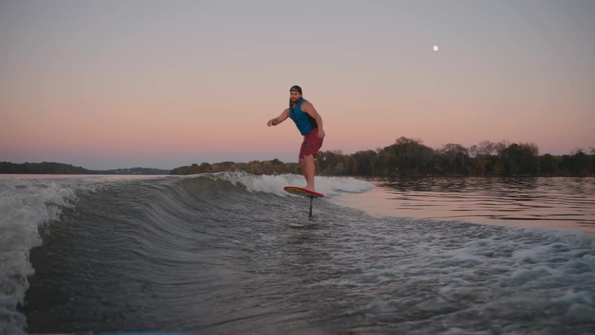 A man foil surfs on a lake at dusk with the moon in a pink orange sky