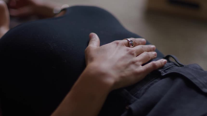 A close view shows a hand with a silver ring resting and gently rubbing the round stomach of a pregnant woman lying down in dark clothing