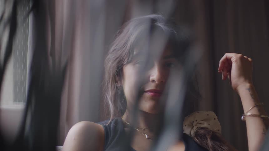 A young woman with long black hair wearing makeup sits indoors beside a window at midday as sunlight streams through