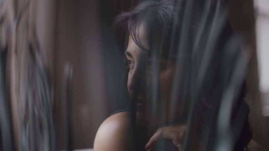 A young woman with long black hair sits indoors smiling gently as she gazes out the window