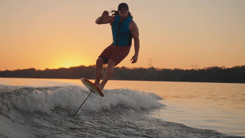 A man in a baseball cap and life vest rides an electric hydrofoil board behind a boat on a calm lake during a warm sunset