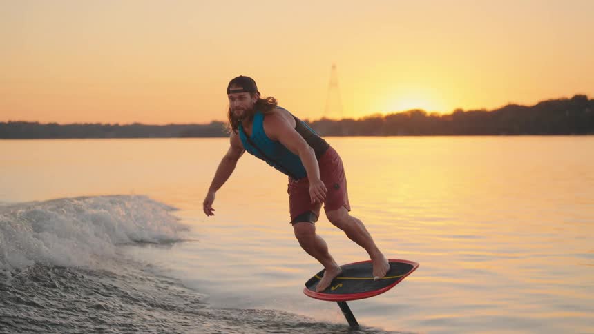 Person on hydrofoil board lifted above calm water at dusk with treelined horizon in the background