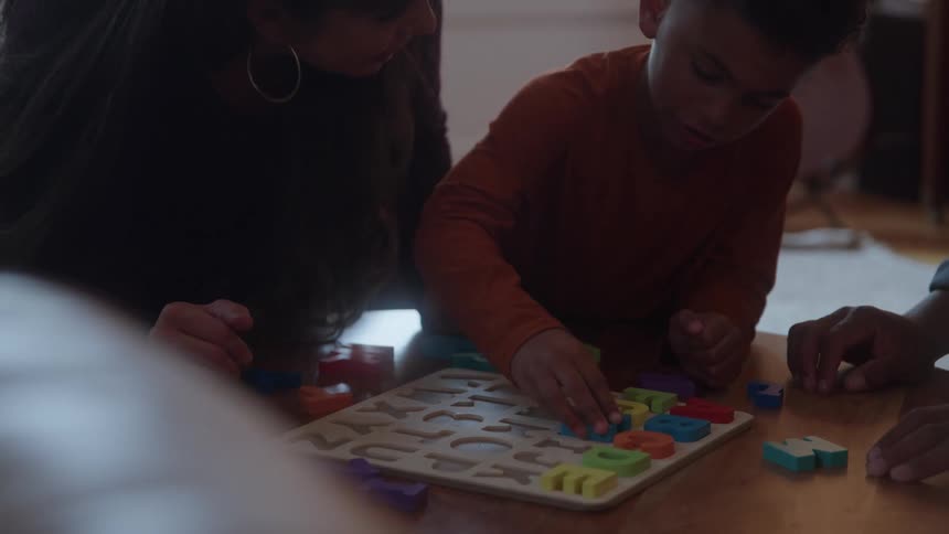 The video shows a group of people including a young boy in an orange shirt gathered around a wooden table they are playing with a brightly colored alphabet puzzle suggesting an educational or family activity