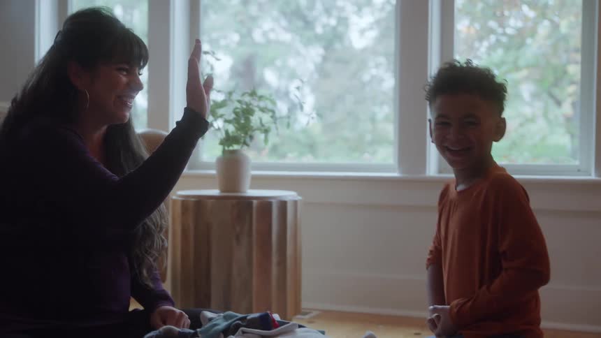 The video shows a pregnant woman sitting on the floor indoors interacting joyfully with a young boy standing opposite herthey are exchanging a high five as the boy laughs and the scene is set in a bright room with large windows showing a lush overc