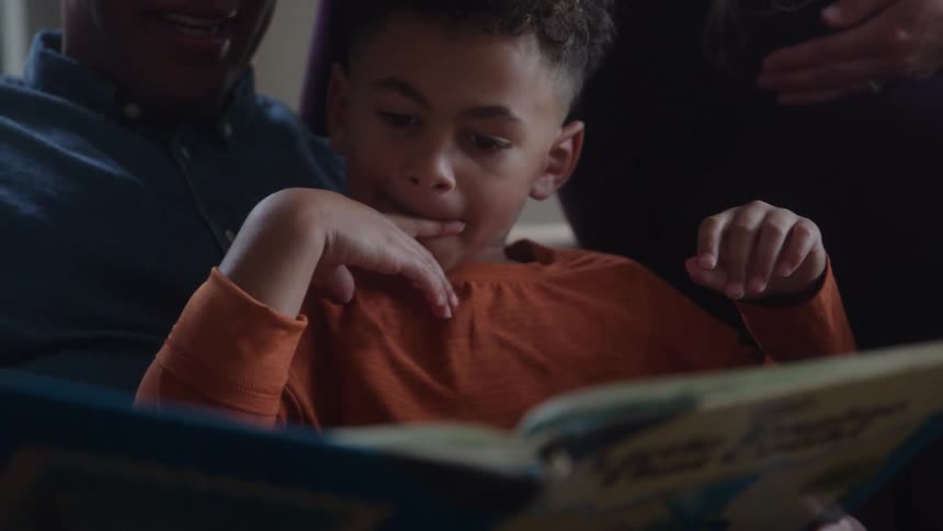 Young boy sitting on the lap of an adult man reading a book and smiling with another person standing next to them indoors during day time