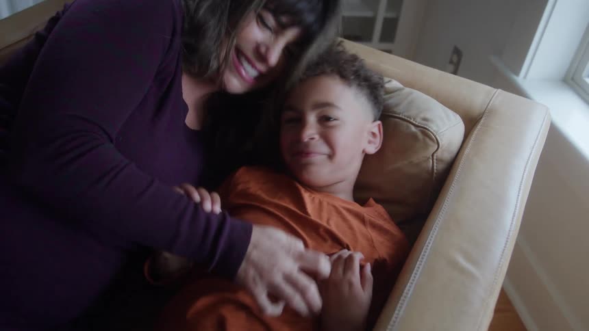 A mother sits on the couch at midday playfully tickling her child as they laugh and enjoy their time together