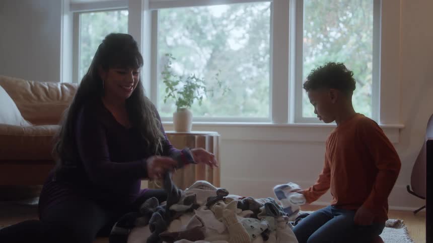Young boy and young adult woman sitting on the floor of a living room laughing and throwing around pieces of clothing while sorting a pile of dirty laundry indoors during day time