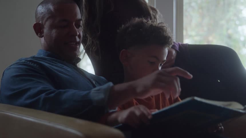This video captures a cozy family moment where a father is sitting on a chair reading a book aloud to his young son who is leaning in to look at the pages a third family member possibly the mother is standing or sitting just behind them resting her h