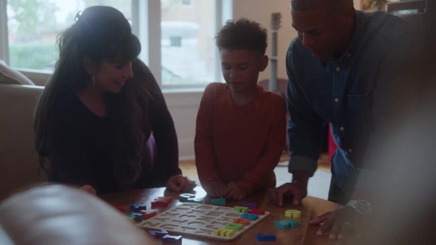 A family of three a father mother and young son all gathers in the living room leaning toward the coffee table as the boy happily plays with letter toys
