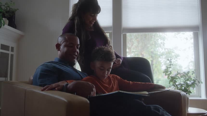 A father carries his son on his lap while sitting in an armchair at midday the mother sits on the arm of the chair watching her son as he plays with a storybook while the father reads aloud