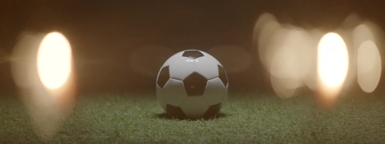 A soccer ball sits centered on a patch of green grass in low light as it comes into sharp focus from a blurred warmtoned backgroundbright circular bokeh lights surround the ball creating an intense atmospheric mood as if under stadium lights at ni
