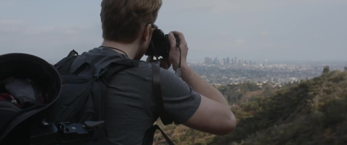 Man wearing backpack takes picture of city from lookout point on smoggy day