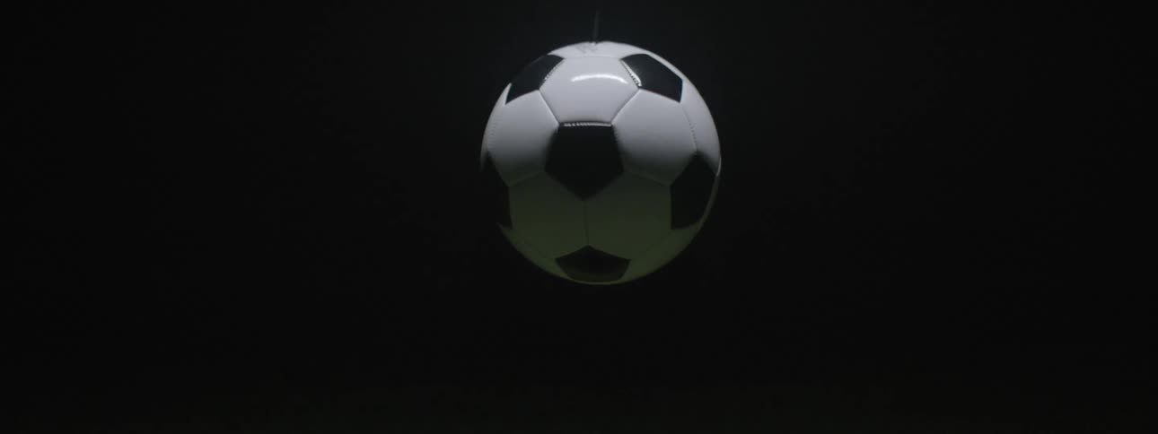 A classic black and white panel soccer ball descending from the dark upper frame until it hangs suspended perfectly centered over a patch of green grass turf