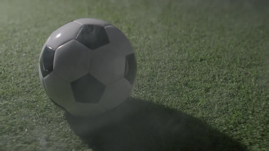 A black and white panel soccer ball sitting static on a patch of green grass turf illuminated by a strong singular light source that casts a long dark shadow across the ground with a subtle mist hovering over the field