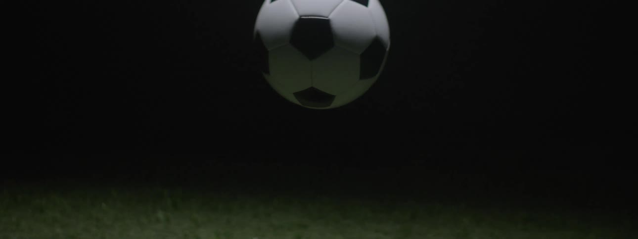 A black and white panel soccer ball that slowly descends from the top of the frame and hangs suspended in midair against a completely dark background with a patch of green turf visible at the very bottom