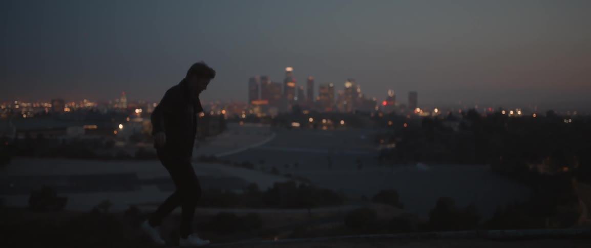 A man balances on a ledge with city skyscrapers in the distance