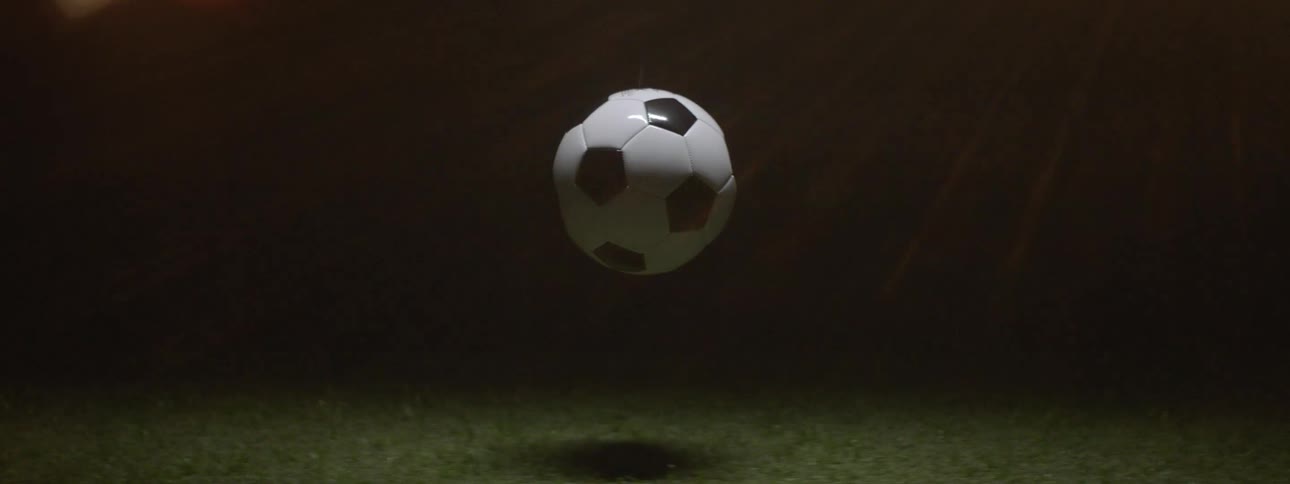Soccer ball hovering above grass with dark background and blurred lights in the distance