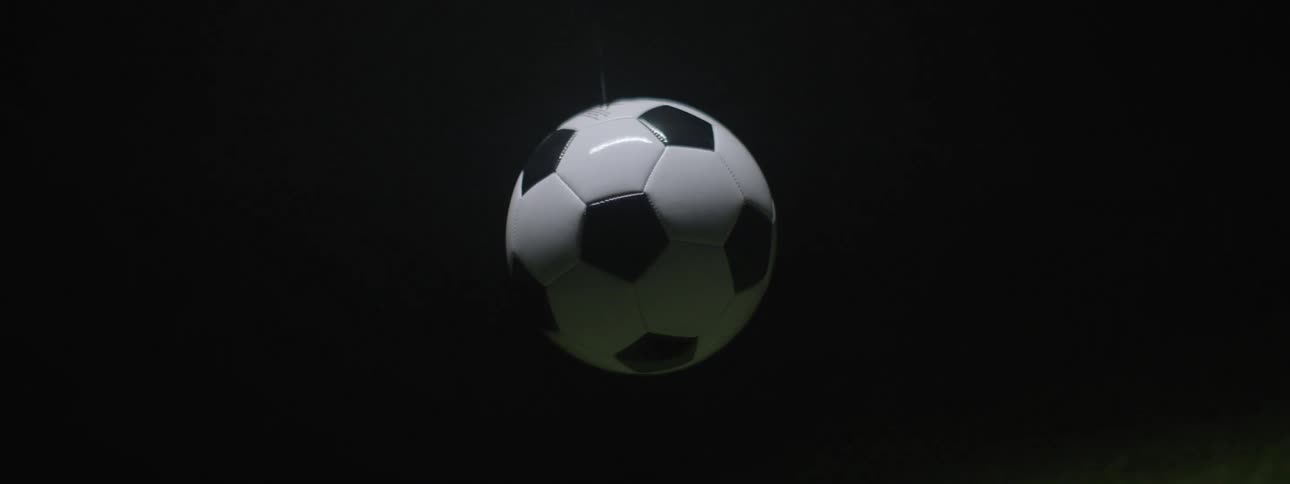 A blackandwhite soccer ball is shown in a studio shot descending from the top of the frame slowly rotating and passing the bottom edge of a patch of illuminated green artificial grass